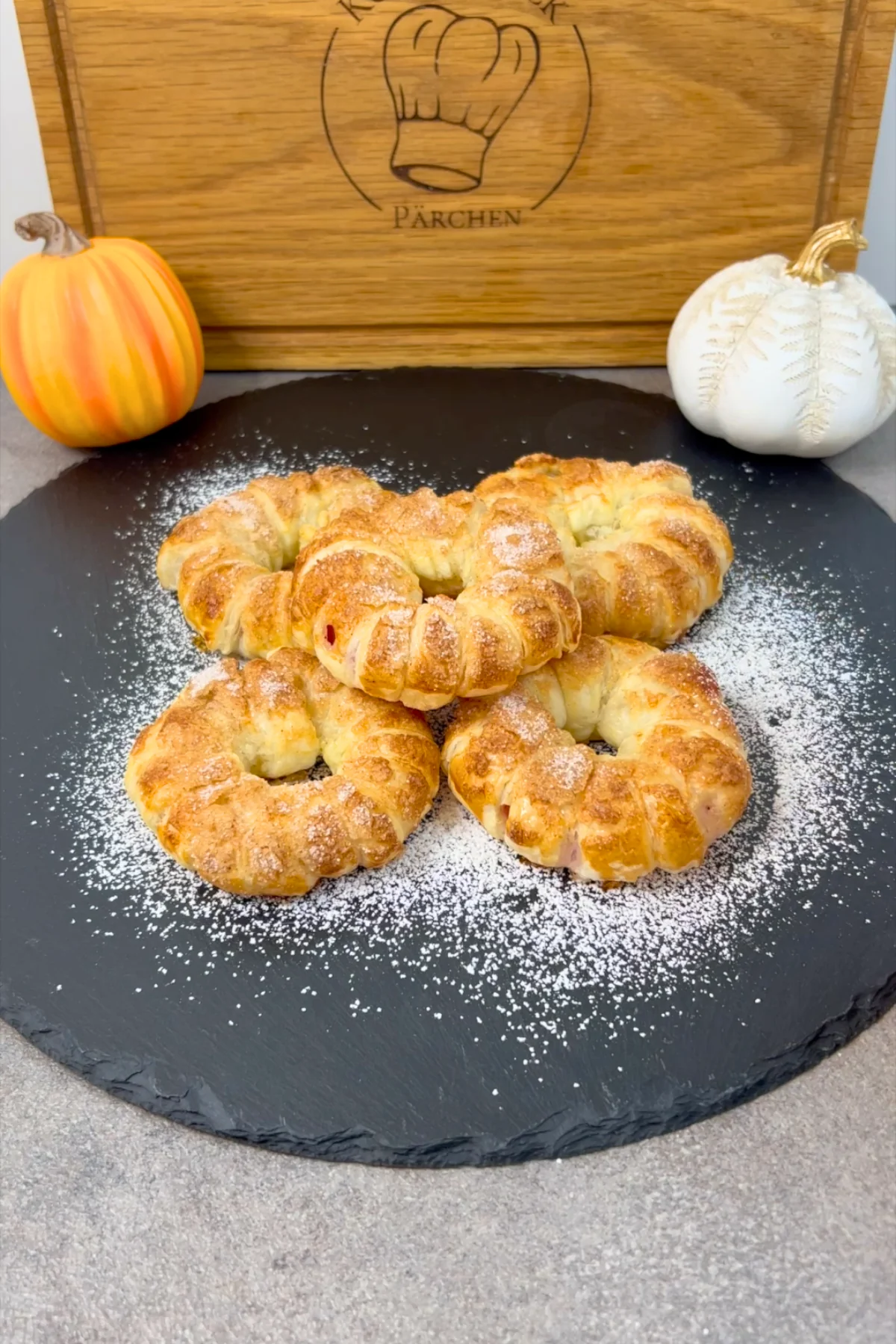 Fertige Apfel-Donuts mit Puderzucker auf Schieferplatte, dekoriert mit herbstlichen Deko-Kürbissen