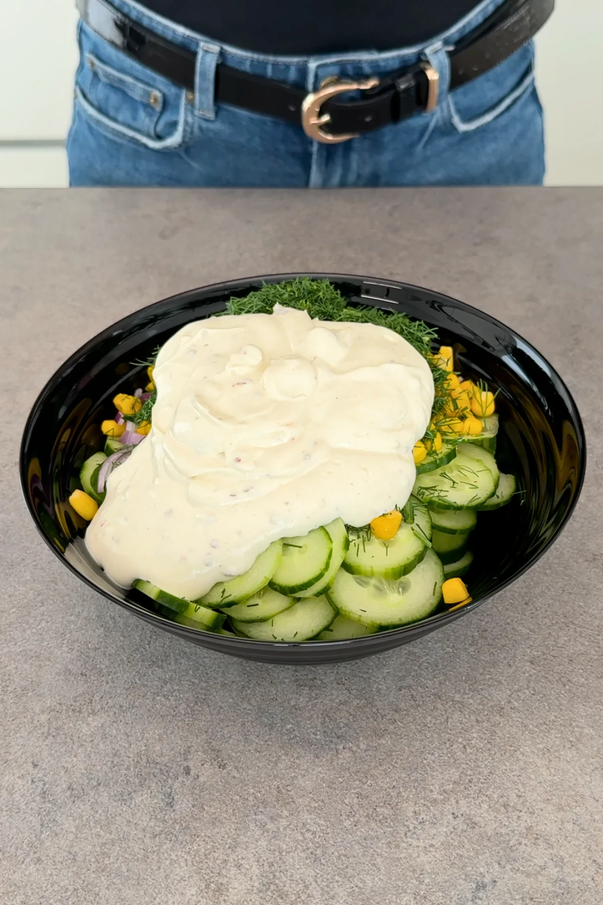 Cremiges Joghurt-Dressing wird über Gurkenscheiben, Mais und Dill in schwarzer Schüssel gegossen