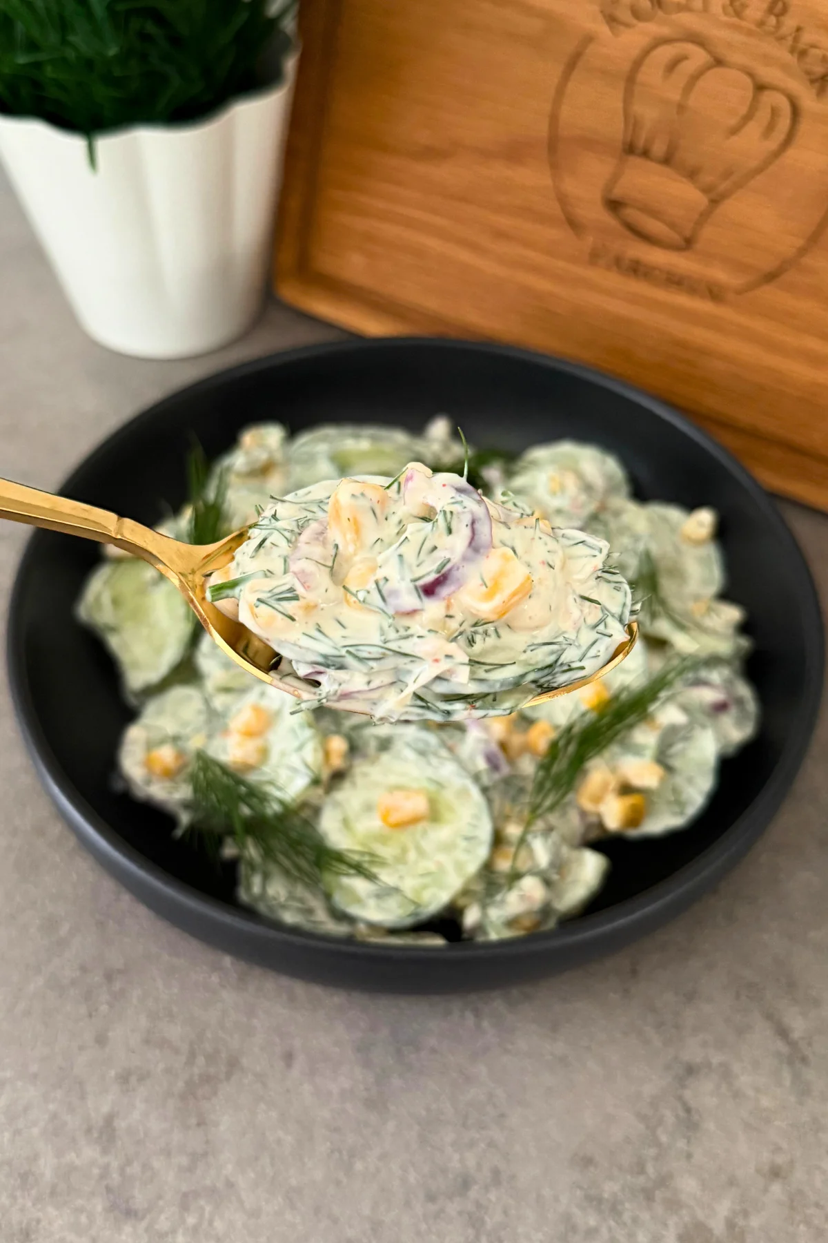 Goldener Löffel hebt eine Portion Gurke-Mais-Salat aus der schwarzen Schüssel, Holzbrett und Pflanze im Hintergrund