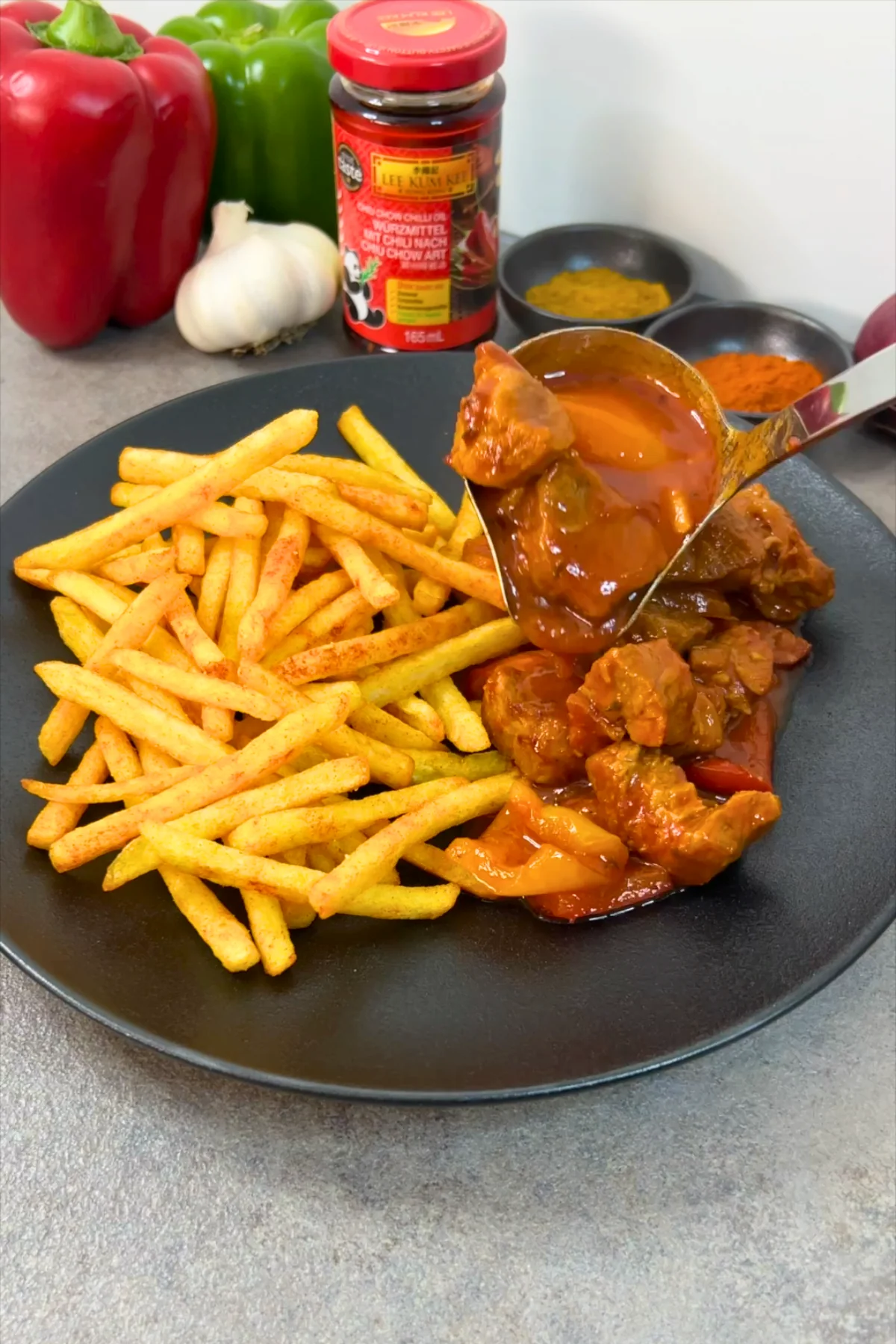 Kelle mit Schaschliktopf über einem Teller mit Pommes, im Hintergrund Gewürze und Chiliöl
