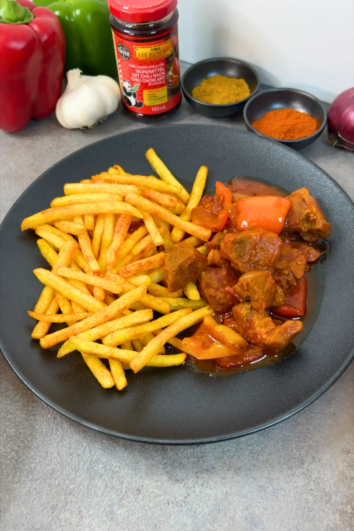 Schaschliktopf mit Pommes auf schwarzem Teller, daneben Paprika, Knoblauch und Chiliöl