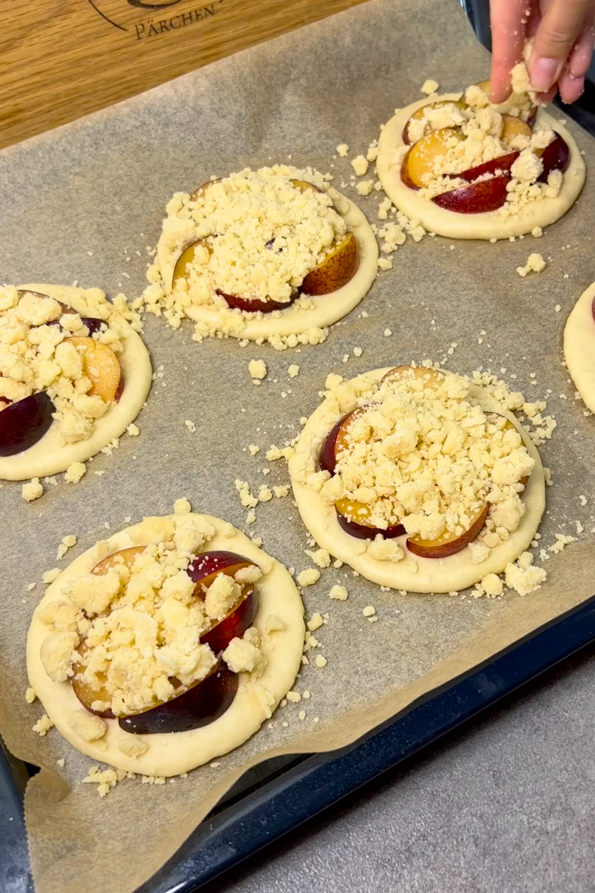 Streusel werden über die mit Pflaumenspalten belegten rohen Teigtaler auf dem Backblech gestreut
