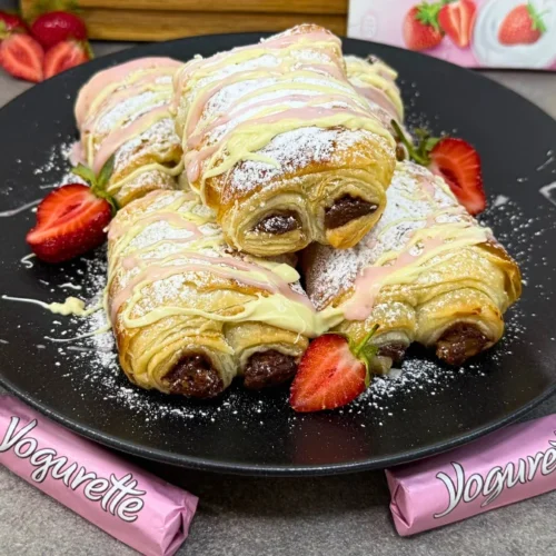 Mehrere Yogurette Croissants mit Puderzucker und weiß-rosa Schokoladenstreifen auf schwarzem Teller