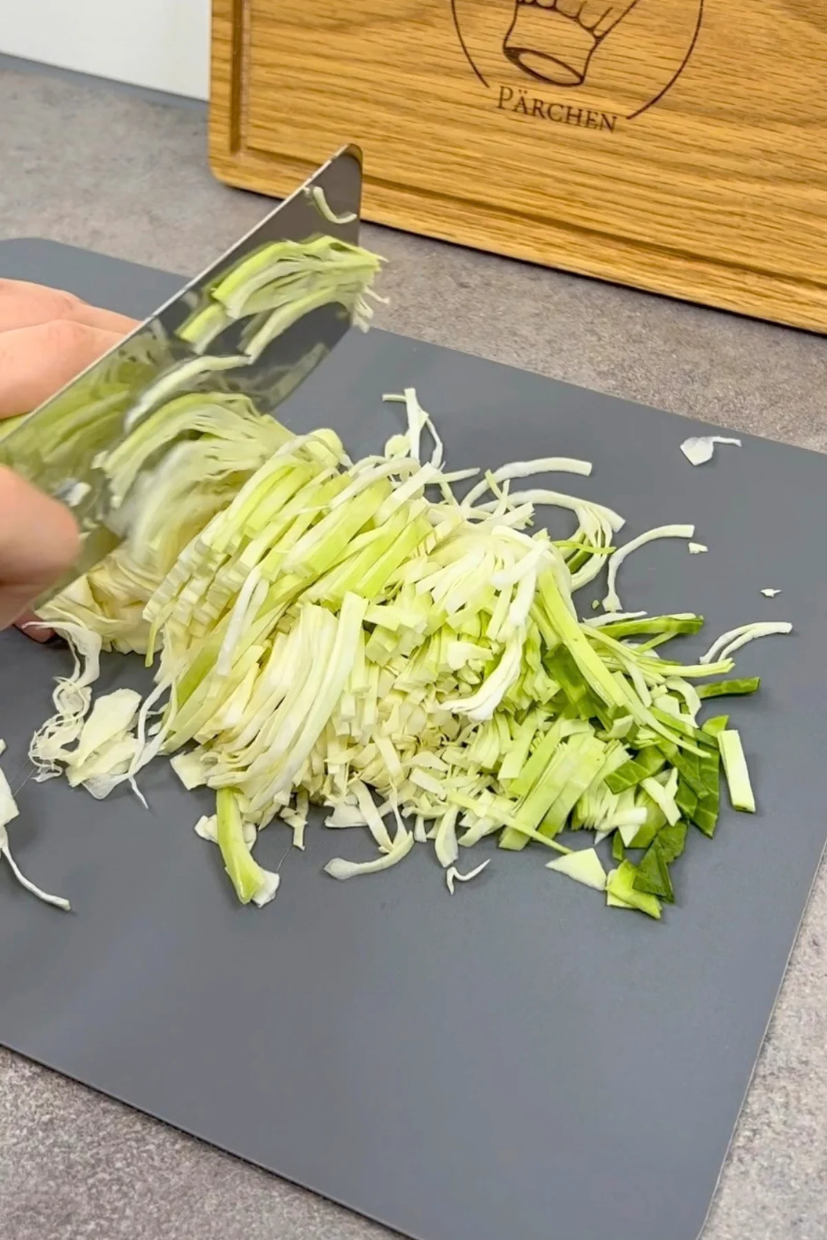 Spitzkraut wird mit dem Messer auf einem grauen Schneidebrett in feine Streifen geschnitten