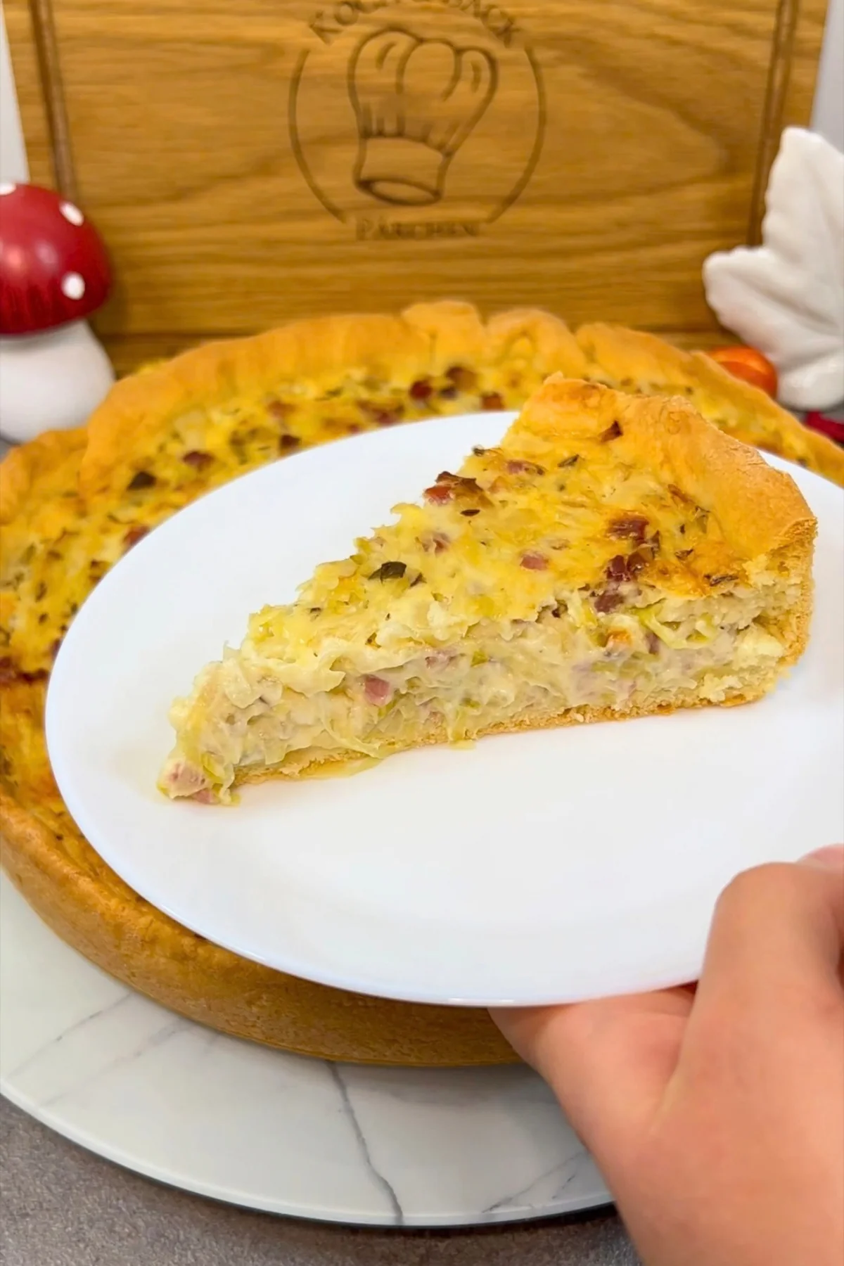 Stück Krautkuchen mit Kraut und Speckwürfeln auf einem weißen Teller vor der runden Backform