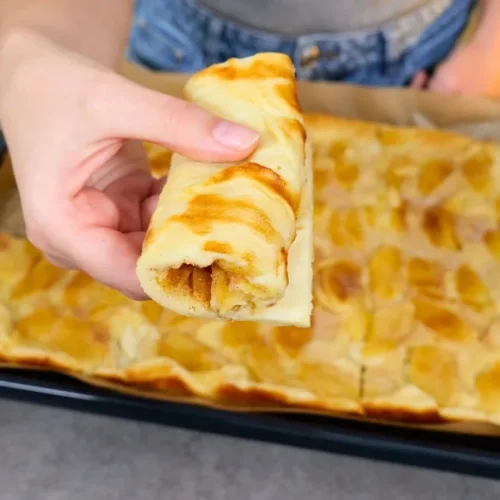 Eine aufgerollte Bahn Apfelpfannkuchen wird über dem gebackenen Blech in der Hand gehalten