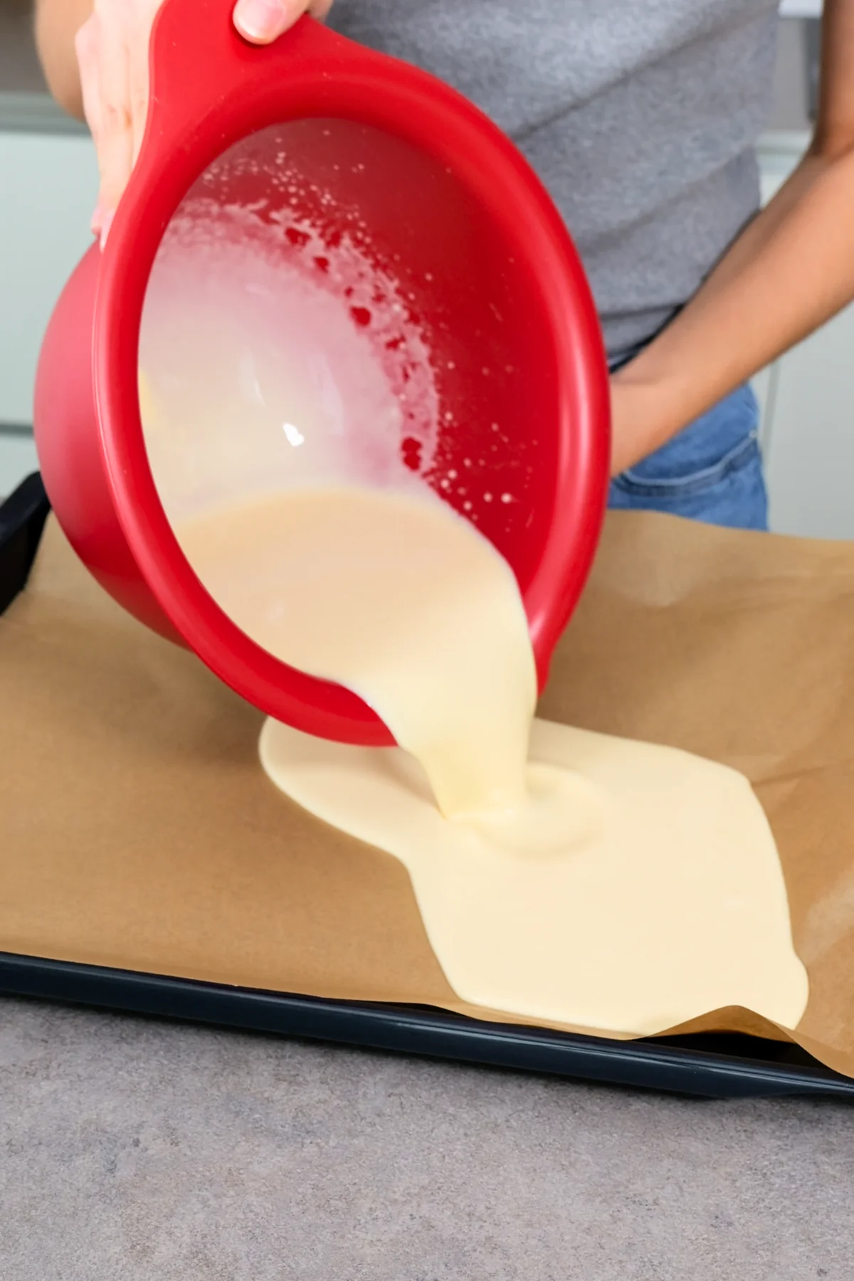 Flüssiger Pfannkuchenteig wird aus einer roten Rührschüssel auf das mit Backpapier belegte Blech gegossen