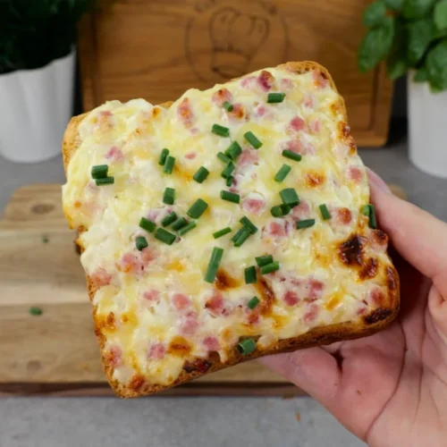 Ein fertiger Flammkuchentoast mit geschmolzenem Käse, Schinkenwürfeln und Schnittlauch wird über einem Holzbrett in der Hand gehalten