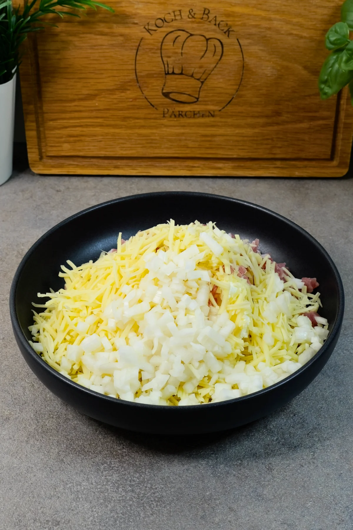 Geriebener Käse und fein gewürfelte Zwiebeln liegen auf den Schinkenwürfeln in der schwarzen Schüssel