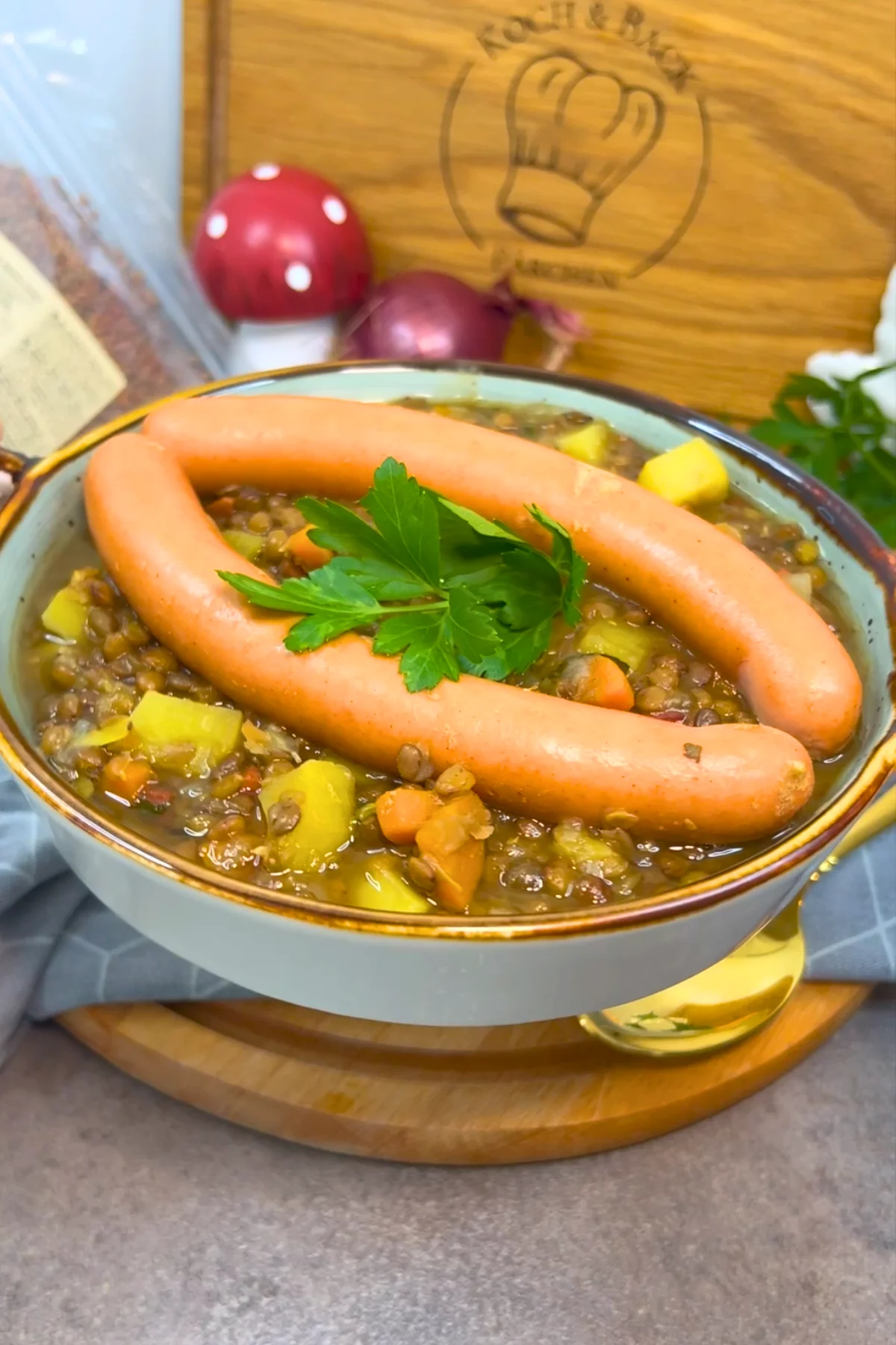 Der angerichtete Linseneintopf auf einem Holzbrett, vor dem Schneidebrett liegen eine rote Zwiebel und ein Deko-Fliegenpilz