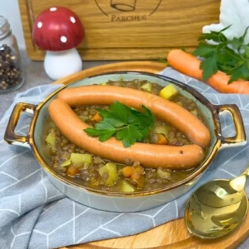 Linseneintopf mit zwei Würstchen und Petersilie in einer runden Schale auf einem Küchentuch, daneben eine Karotte, ein Pfefferglas und ein goldener Löffel