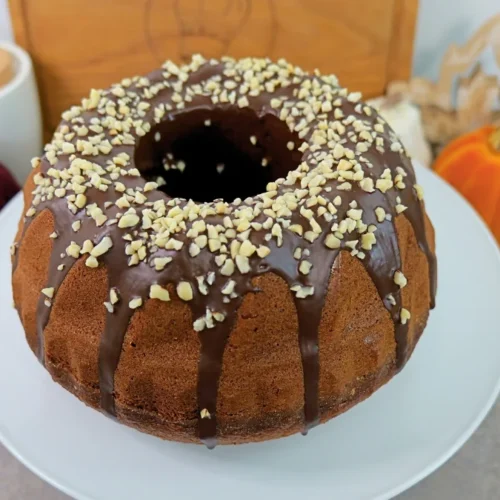 Ganzer Nusskuchen Gugelhupf mit Schokoglasur und gehackten Mandeln auf einem weißen Teller, daneben zwei Deko-Kürbisse