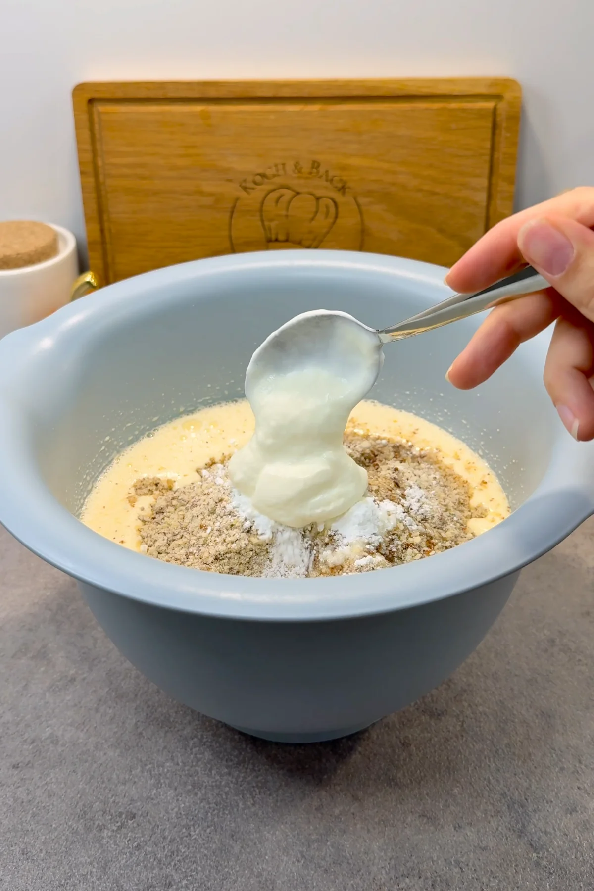 Ein Löffel Naturjoghurt wird zum Schluss über die Nuss-Mehl-Mischung in der blauen Rührschüssel gegeben