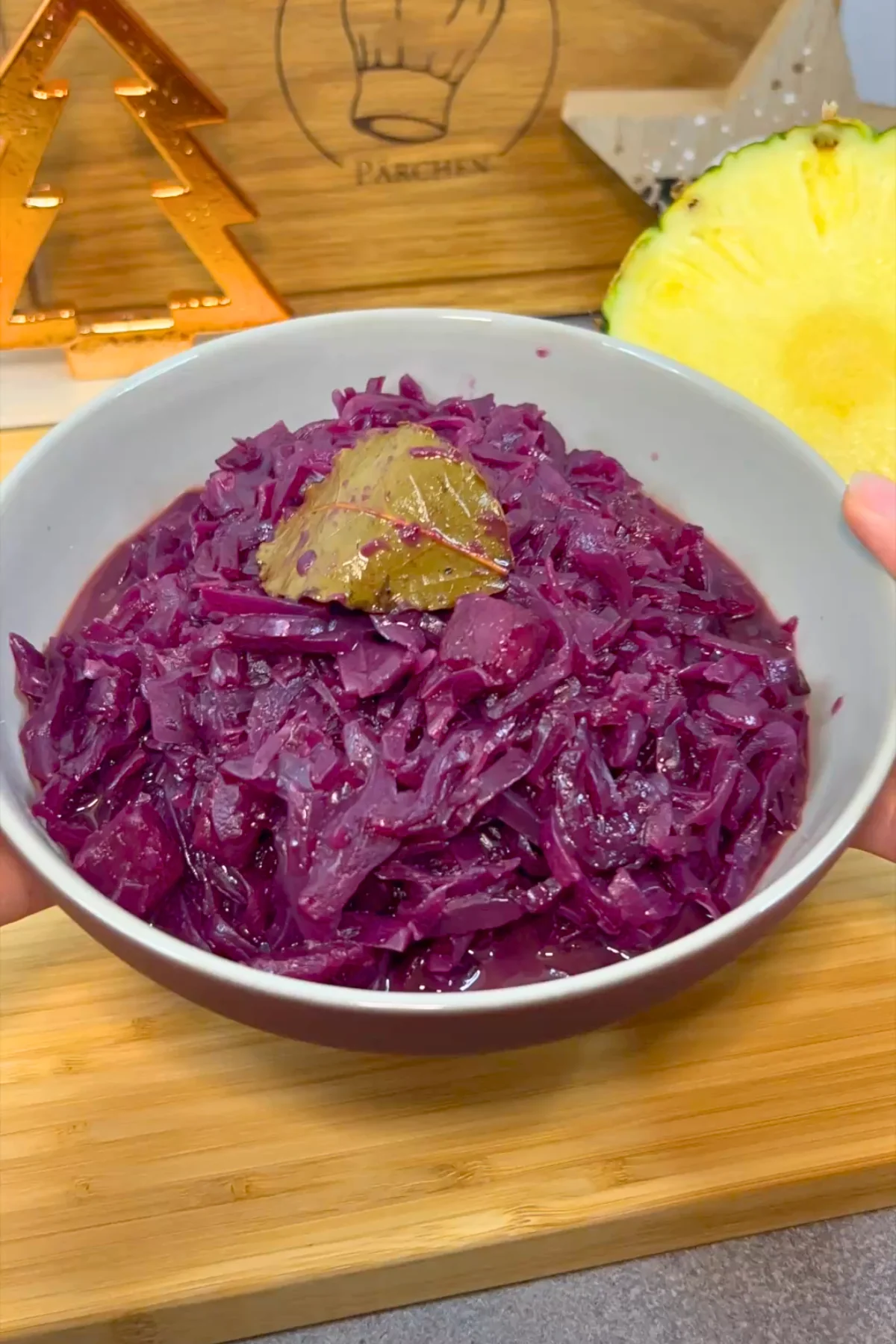 Die Schale mit fertigem Rotkohl und Lorbeerblatt wird vor einer halben Ananas in der Hand gehalten