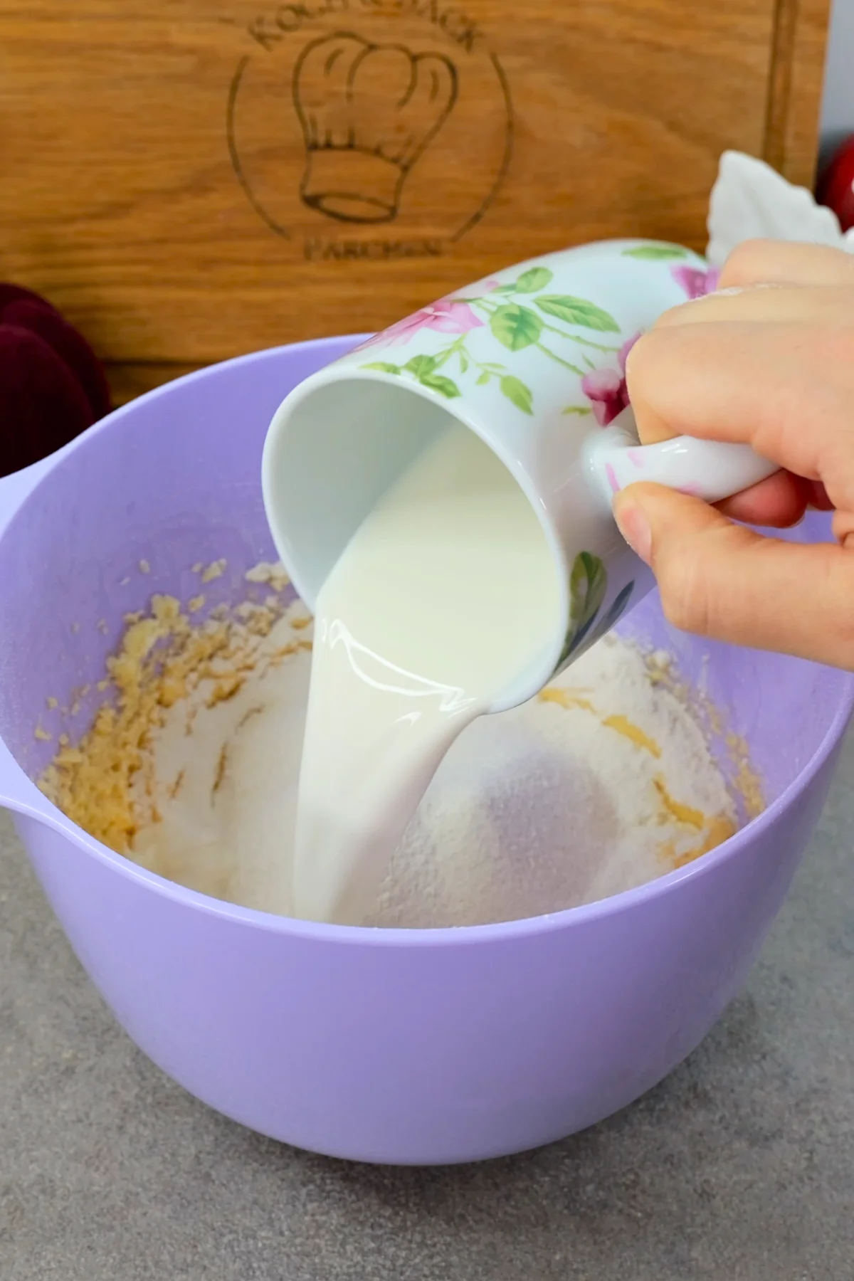 Milch wird aus einem Blumenkrug über das gesiebte Mehl in der lila Rührschüssel gegossen