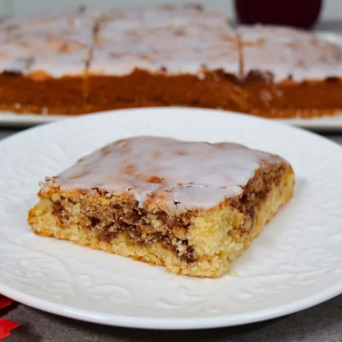 Ein Stück Zimtschneckenkuchen mit Zuckerguss liegt auf einem weißen Teller, dahinter der angeschnittene Kuchen auf einer Platte