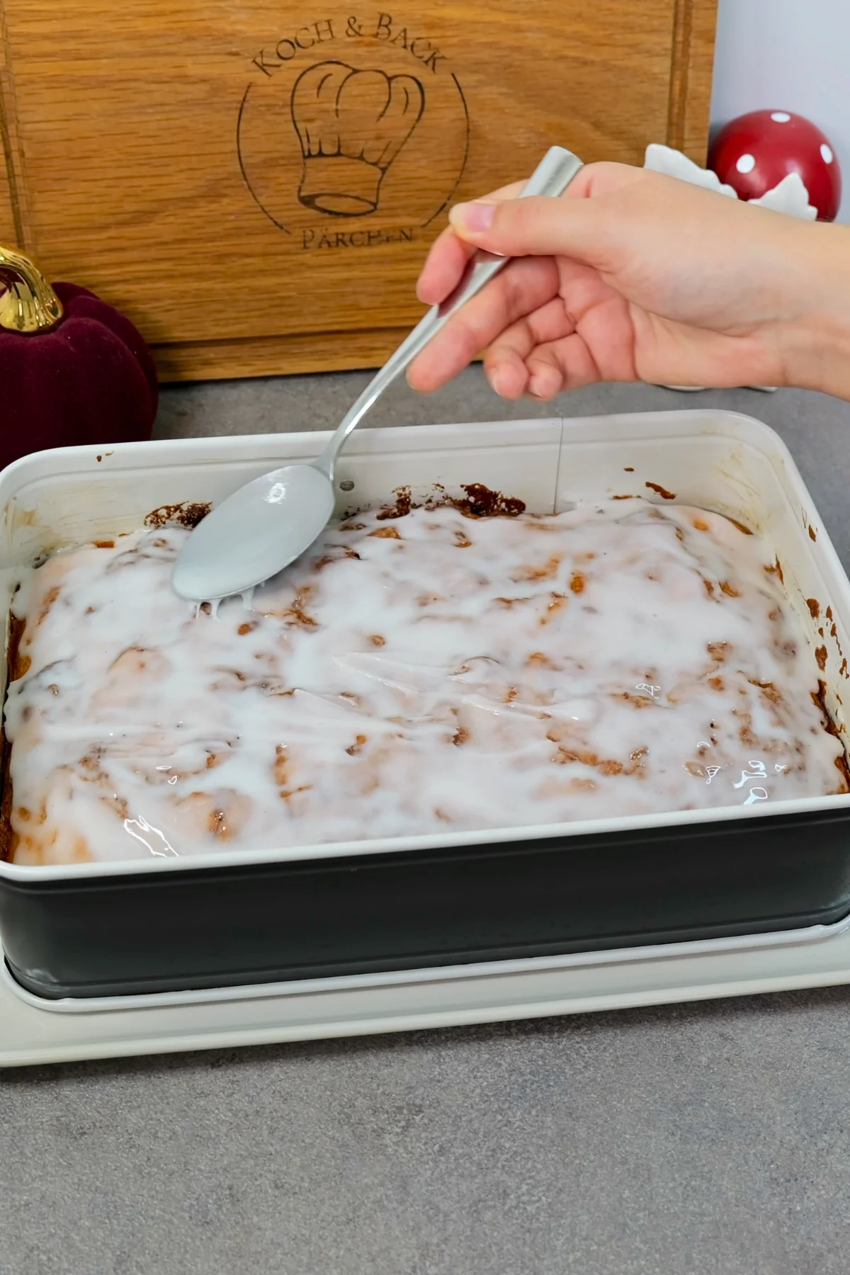 Weißer Zuckerguss wird mit einem Löffel auf dem noch in der Form liegenden Zimtschneckenkuchen glatt gestrichen